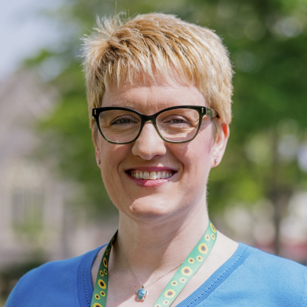 Lisa, a middle-aged woman with short blonde hair is wearing glasses, a sunflower lanyard an a Mediterranean blue jumper. She is smiling and pointing up to her left.